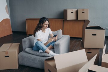 Girl uses phone app, orders moving company, shopping online for new home, sitting with carton boxes