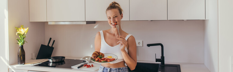 Positive blonde woman eating cherry tomato and holding breakfast in kitchen, banner.
