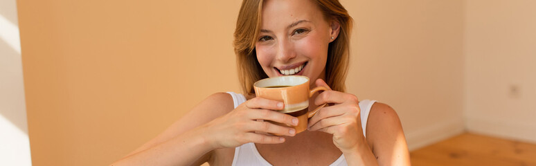 Positive blonde woman holding cup of coffee and looking at camera at home, banner.