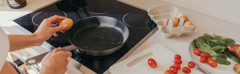 Cropped view of woman holding egg near frying pan and cherry tomatoes in kitchen, banner.