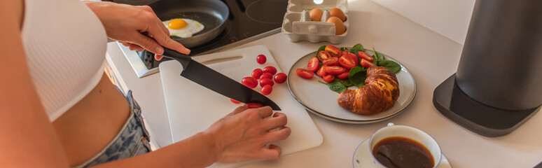 Cropped view of woman cutting cherry tomatoes near coffee and croissant in kitchen, banner.