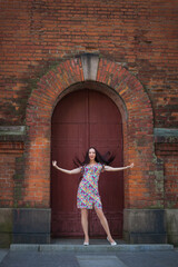 Fototapeta na wymiar A beautiful slender smiling woman with long dark hair, in a light summer dress with a floral pattern, stands at the door in the form of an arch of an old red brick building.