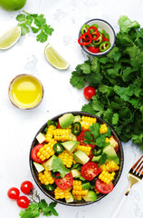 Mexican spicy salad with corn, avocado, jalapeno peppers, cherry tomatoes and cilantro. White kitchen table background, top view