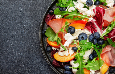 Fresh salad with arugula, radicchio, sweet peaches, smoked pork ham, feta cheese and blueberries. Black stone table background, top view, close up