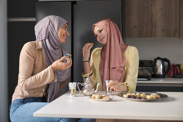 Two young muslim women talking, drinking tea and eating arabic sweets at the kitchen table.