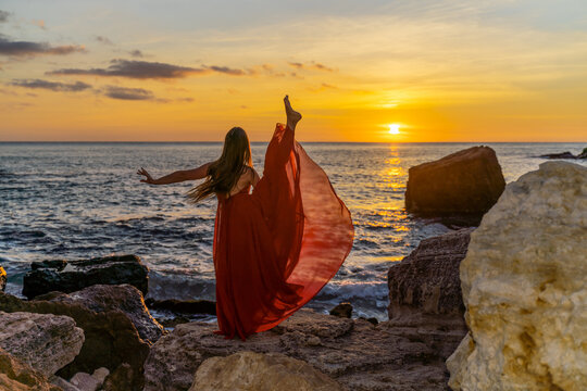 a dancing woman in a red flying dress on the ocean or on the sea beach against the backdrop of the sunset sky.