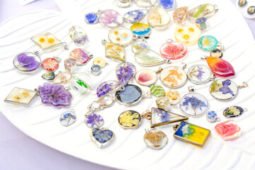 Epoxy resin jewelry - many handmade pendants with real flowers and plants inside on white stand. Making exclusive jewellery at home - obrazy, fototapety, plakaty