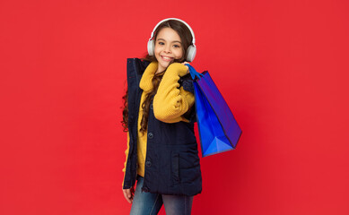 happy child in winter clothes and headphones hold shopping bag on red background, sale
