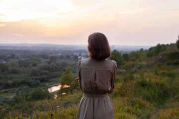 Fototapeta na wymiar Beautiful young smiling girl in a long brown dress stands along the lawn. Happy woman walks at sunset on a hill overlooking the river. Concept of having rest in park during summer holidays or weekends