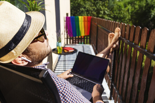 Male person in sun hat and sunglasses listening to music with headphones and using his laptop on the terrace of hotel