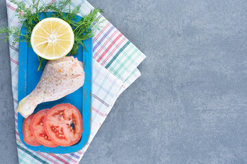 Uncooked chicken leg, tomato and lemon slice on blue plate