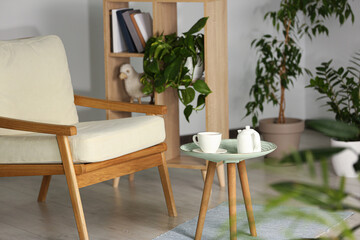 Stylish armchair, side table with cup and teapot near beautiful houseplants. Interior design