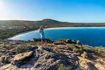 a long-haired girl in a dress stands at the top of a mountain admiring the view of a vast bay of white sand and turquoise water in cape le grand national park