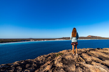 a long-haired girl in a dress stands at the top of a mountain admiring the view of a vast bay of...