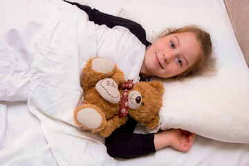 little girl lies in bed with a teddy bear and smiles