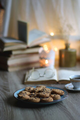 Plate of chocolate chip cookies, stack of vintage books, reading glasses, cup of tea or coffee, lit candle and fairy lights. Hygge at home. Selective focus.