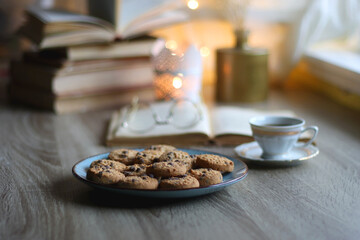 Plate of chocolate chip cookies, stack of vintage books, reading glasses, cup of tea or coffee, lit candle and fairy lights. Hygge at home. Selective focus.