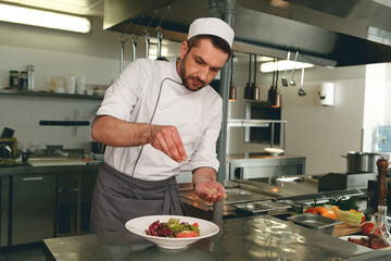 Chef sprinkles salt to salad of fresh vegetables in kitchen of restaurant. Tasty and healthy food