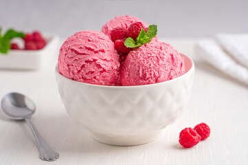 Scoops of sweet refreshing homemade pink berry ice cream or organic sorbet made of ripe juicy raspberries served in bowl decorated with mint leaf on white wooden table with metal spoon and towel