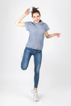 Studio portrait of gorgeous brunette woman with beautiful body shape wearing blue sweater without sleeves, jeans and sport shoes jumping in air. Image contains motion blur and noise
