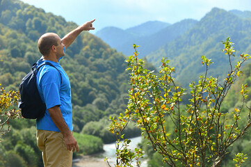 a male tourist in a blue T-shirt and with a backpack stands on a high slope of a mountain and points forward with his hand