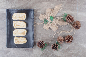 Cake roll slices on a black platter next to a festive decoration made of pine cones on marble background
