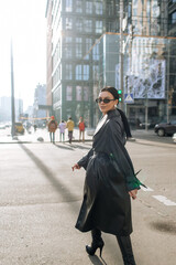 Girl on the street in a black coat. Stylish girl in a black raincoat on the background of a high glass house