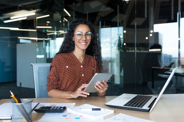 Happy and successful hispanic woman working inside modern office building, business woman using tablet computer smiling and looking at camera worker doing paperwork. - obrazy, fototapety, plakaty