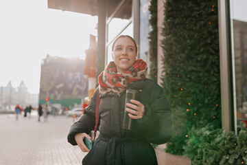 Smiling happy european woman with wonderful smile wearing black jacket and bright scarf enjoys coffee while enjoys walking in cold sunny day. 