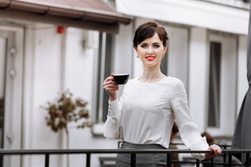 Young beautiful woman holding a cup of coffee or tea in cafe, restaurant. Brunette girl is drinking hot drinks, looking in camera outdoors on terrace and having lunch break during work. copy space