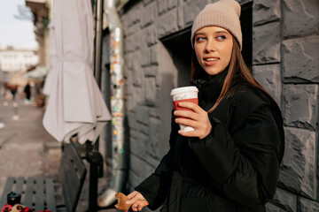 Beautiful stylish european woman in jacket and beige knitted cap drinking coffee on sunny street in...