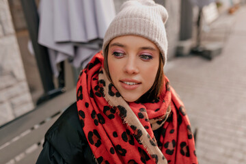 Close up outdoor photo of european pretty woman with bright eyes wearing cap and scarf resting in the city in winter sunny day. 