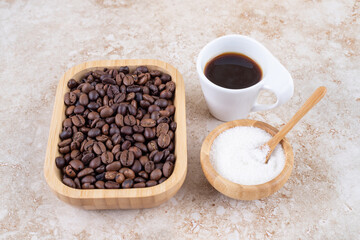 Pile of coffee beans in a wooden platter next to a small bowl of sugar and a cup of coffee on marble background