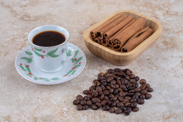 Bundles of cinnamon sticks and coffee beans next to a cup of coffee on marble background