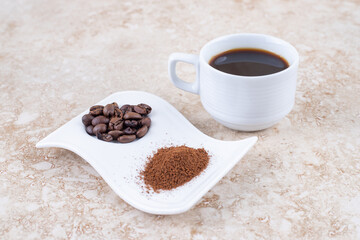 A cup of brewed coffee next to coffee beans and ground coffee powder on a fancy platter on marble background