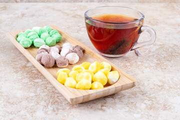 A cup of tea and assorted candies bundled in a small wooden tray on marble background