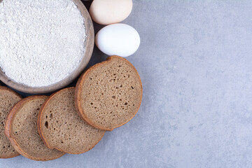 Sliced black bread and eggs around a bowl of flour on marble background