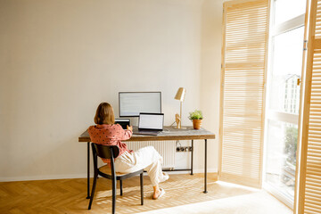 Woman works on computers while sitting by a cozy workplace in sunny living room at home. View from the backside. Concept of remote work from home office. Mockup image