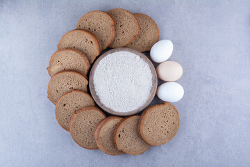 Circle of sliced black bread and eggs around a bowl of flour on marble background