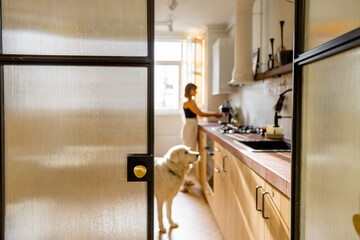 Woman makes coffee while standing with her dog in kitchen. View through the glass door. Domestic lifestyle concept