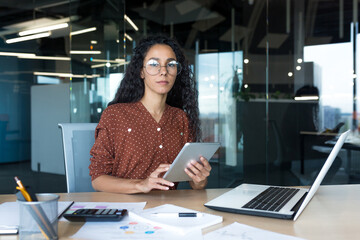 Thinking businesswoman with tablet computer looking seriously at camera, Hispanic woman working...