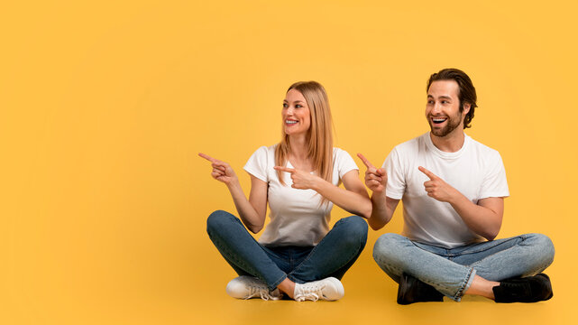Great ad and offer. Smiling millennial caucasian couple in white t-shirts point to empty space, sit on floor