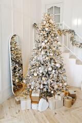 A large Christmas tree decorated with balls and garlands and boxes with gifts on the floor in the interior of a bright living room with a wooden staircase leading to the second floor. Nobody