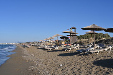 Fototapeta na wymiar Sandy beach of a holiday resort with plastic deck chairs and straw beach umbrellas. Among them there is a tower for lifeguard. Copy space is available. Cloudless sky is on the background.