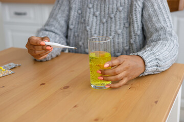 African American woman with a cold measures her body temperature and drinks soluble pills or vitamins, A tired sick woman at home takes pills for sore throat and viruses. treatment of influenza and