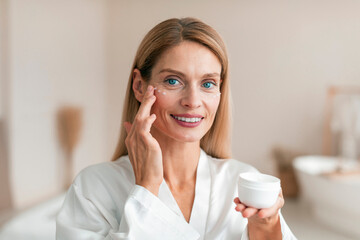 Beautiful middle aged woman in silky bathrobe using moisturizing eye cream, enjoying face care...