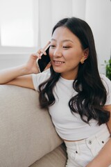 A young Asian woman sits at home on the couch relaxing and talking on the phone on her day off. Lifestyle without work in a comfortable home