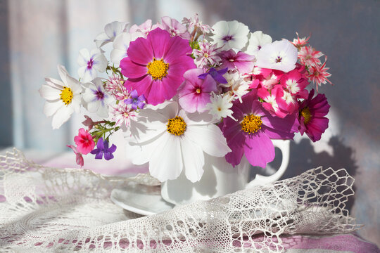 Bouquet of white and pink cosmos flowers, phloxes, bluebells in a white cup on the table, sunlight.