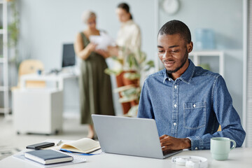 Portrait of young black man using laptop at office workplace in minimal interior, copy space