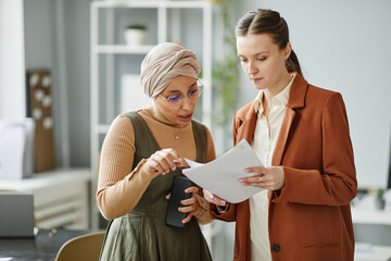 Waist up portrait of two young business women Middle Eastern and Caucasian standing in office...
